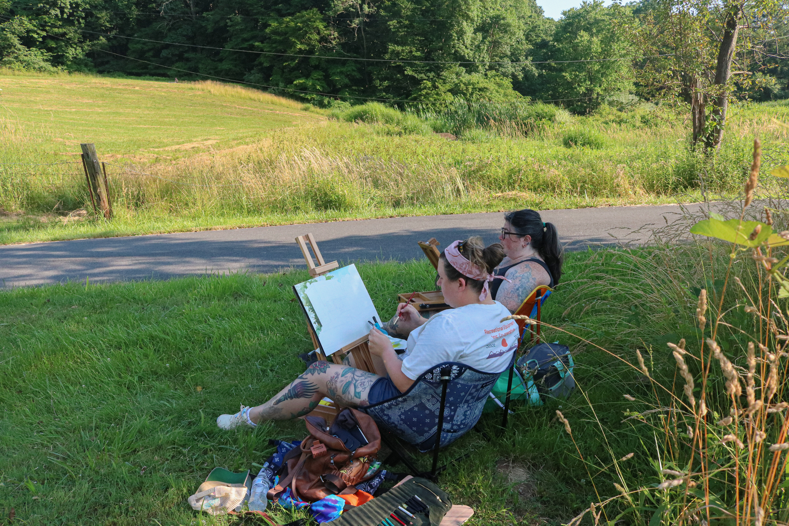 Two women sitting on lawn chairs paint on canvas in an open field.
