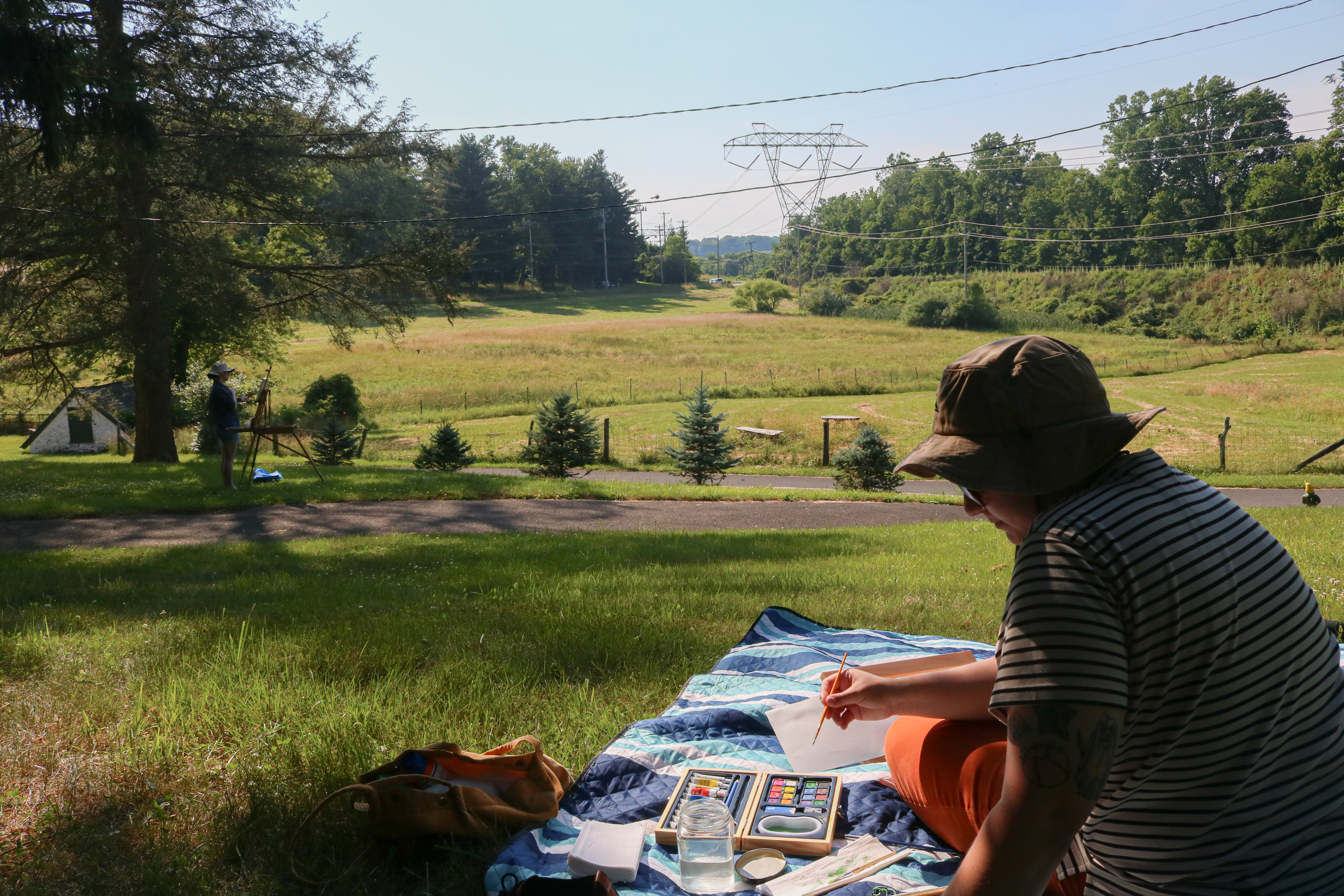 A person sits on a blanket painting in an open area overlooking a small valley.