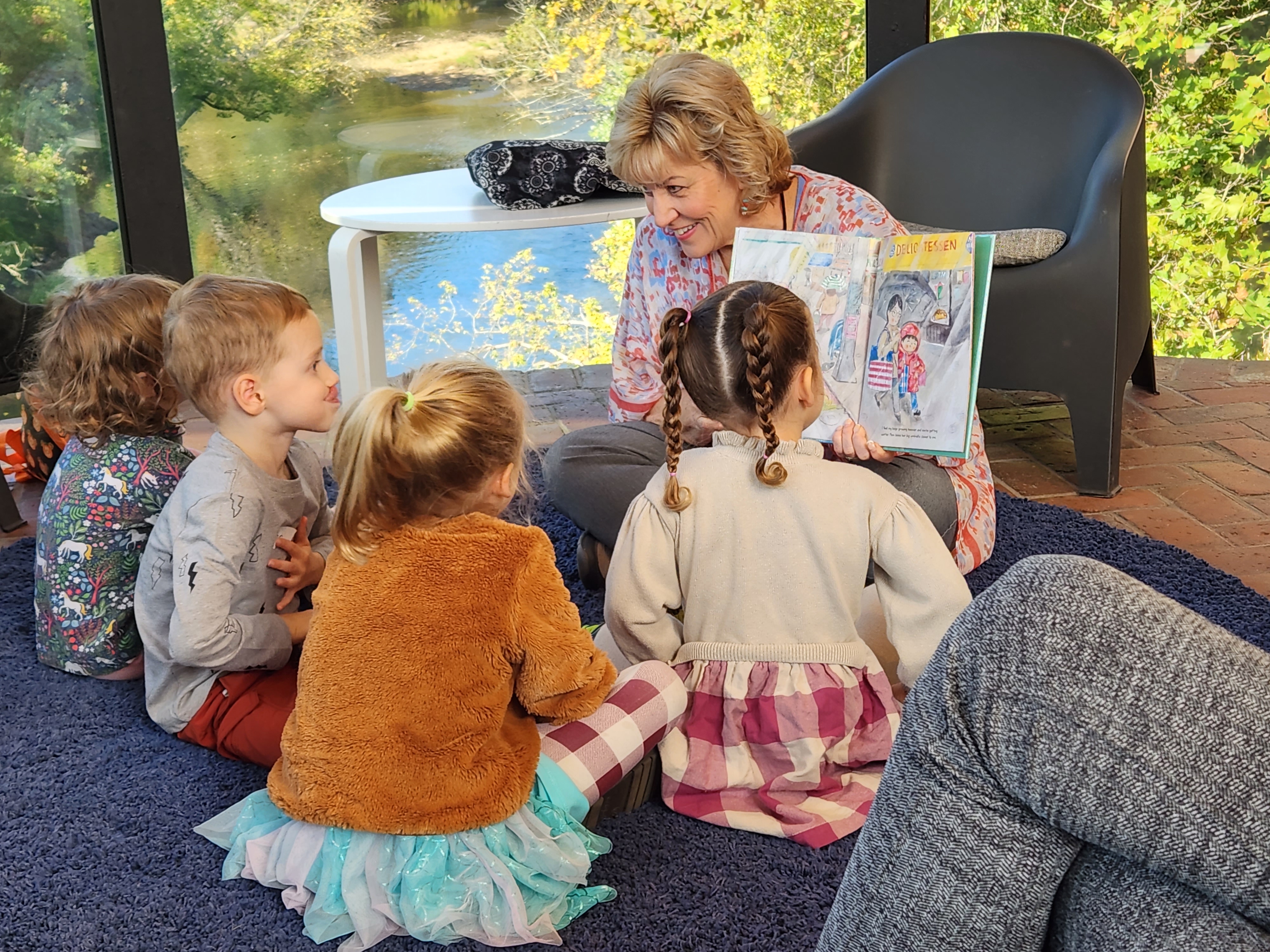 An older women sits on a chair reading a book to a group of younger children who are sitting on the carpeted floor surrounding her.