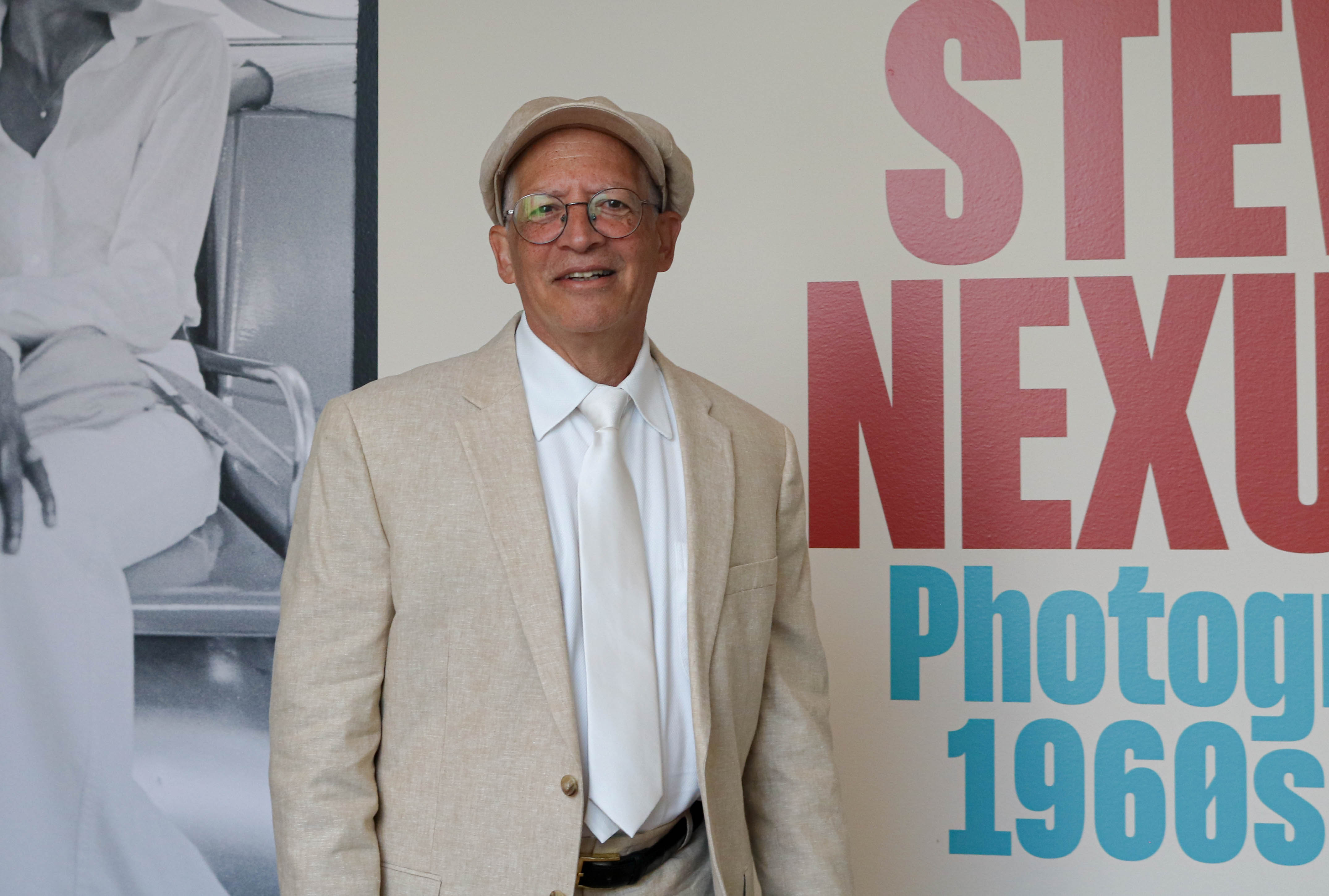 A man in a khaki suit and hat stands against a colorful wall of text, posing for a portrait.