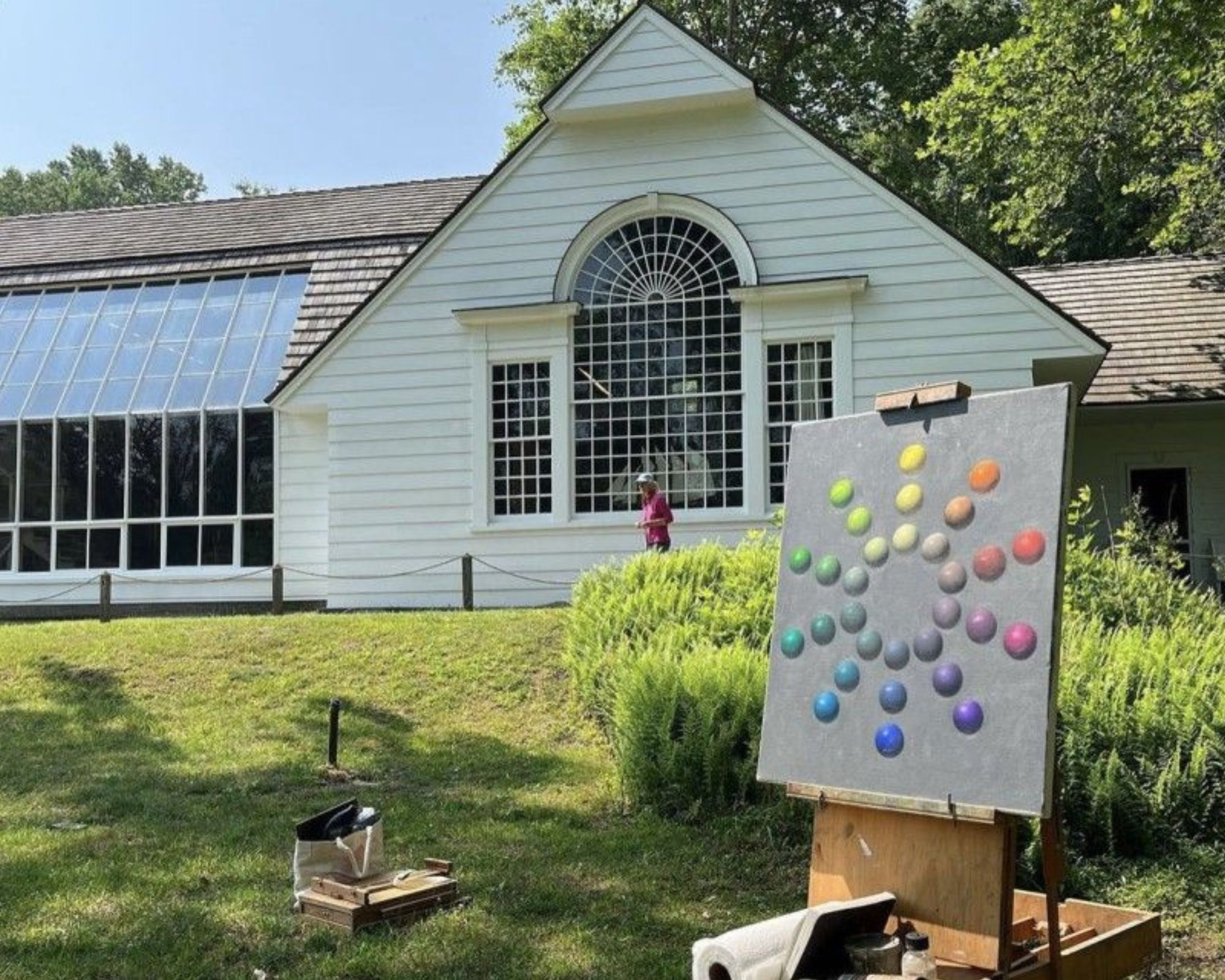 A large house on a summer day with glass windows sits in the background behind a easel with painting supplies.