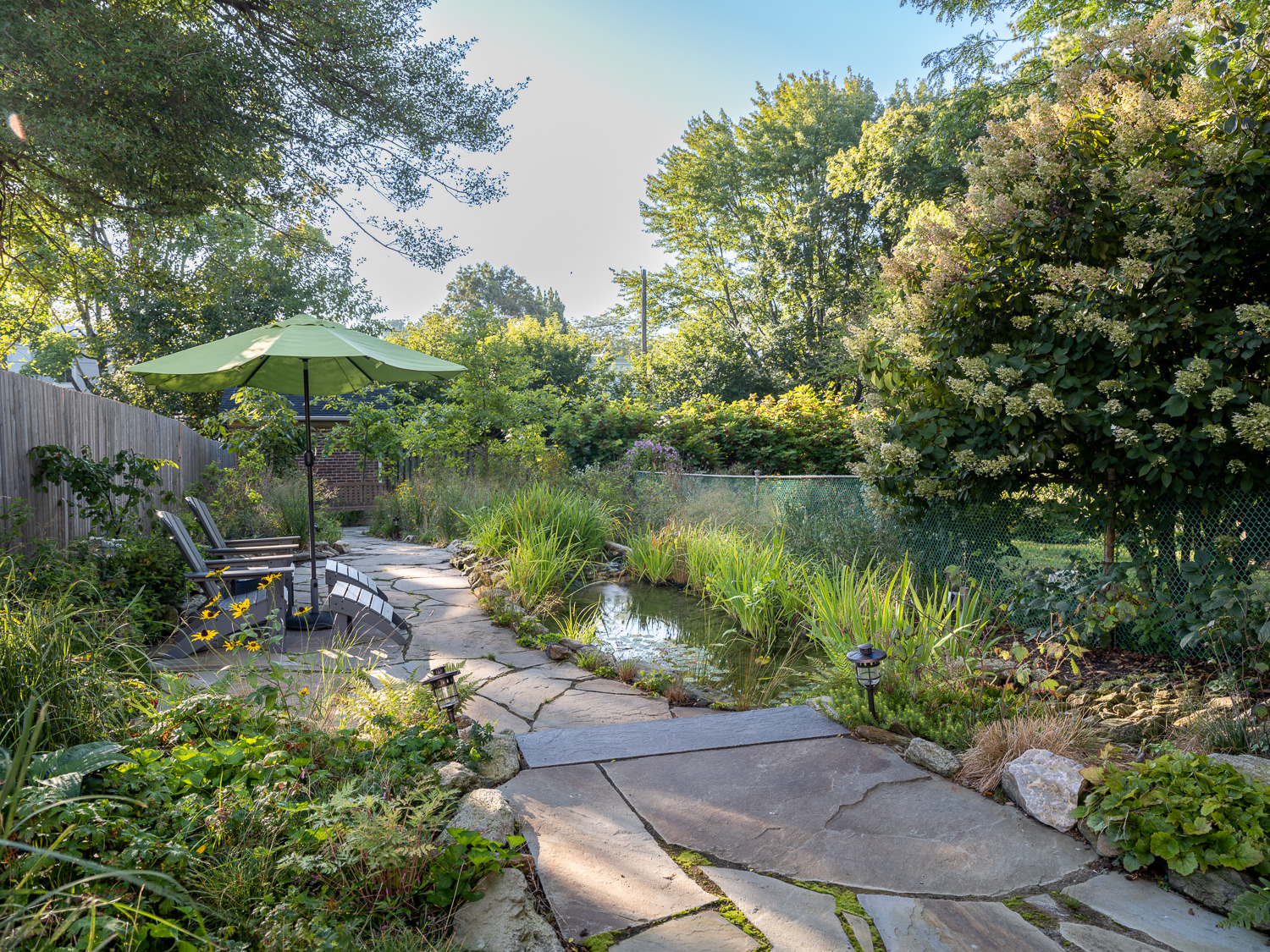 A yard with a stone walkway next to a pond, and two chairs next to a green umbrella