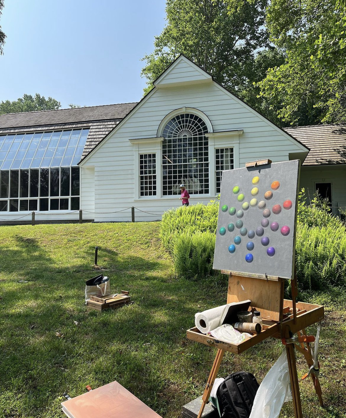 Easel wit a painting on it, sitting in a green garden in front of a white house.