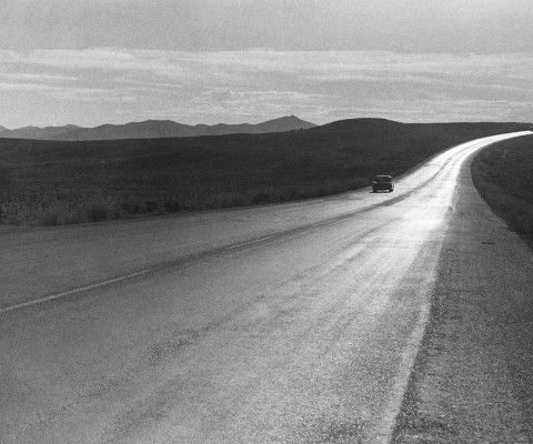 A black and white shot of an open road from the perspective of the side of the road with mountains in the distance.