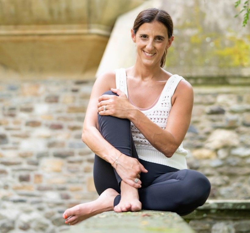 a woman wearing yoga clothes sits with legs folded while smiling at the camera