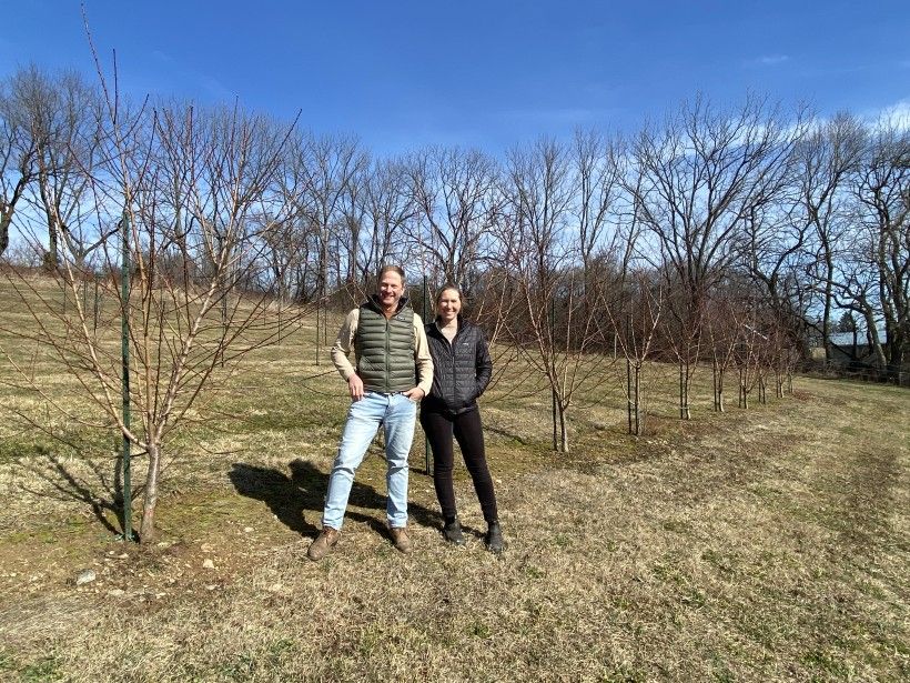 A couple stands to pose for a photo on an open field of their family property.