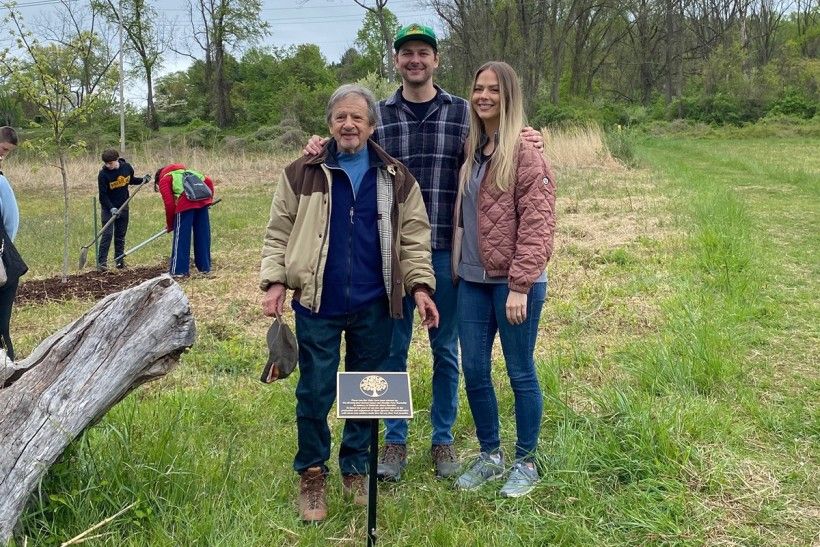 three people stand in front of a plaque during a tree planting memorial