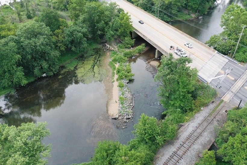 aerial photograph of a dam, breached on one side, in a waterway flowing under a bridge