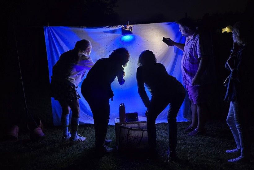 Four figures stand close to a white sheet outside at night, under a UV light, looking for moths