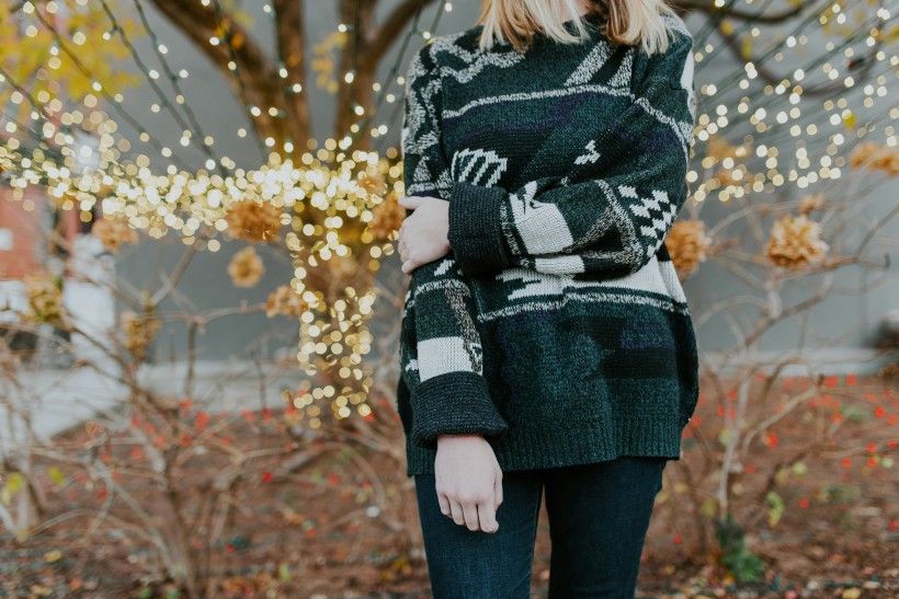 photo of a woman standing with arm folded wearing a sweater in front of a tree wrapped in holiday lights