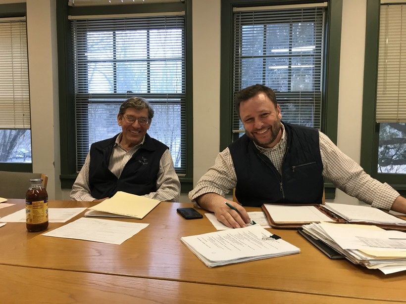 David Shields and Grant DeCosta smile at the camera while sitting behind a conference table