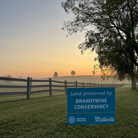 A sign in the foreground that reads "Land Preserved by Brandywine Conservancy" with a sunset over a meadow in the background.